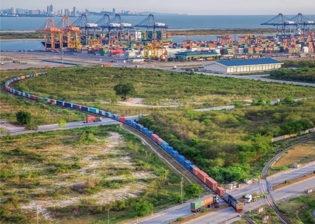Vue aérienne d’un port à conteneurs avec grues portuaires, terminal maritime et train de fret transportant des conteneurs reliant le port aux infrastructures terrestres, illustrant l’intermodalité et la chaîne logistique.